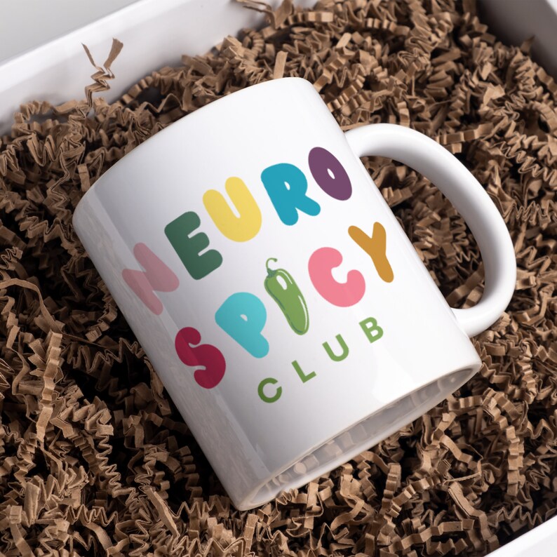 May include: White ceramic mug with a colorful design that says "Neuro Spicy Club". The mug is sitting in brown packing peanuts.