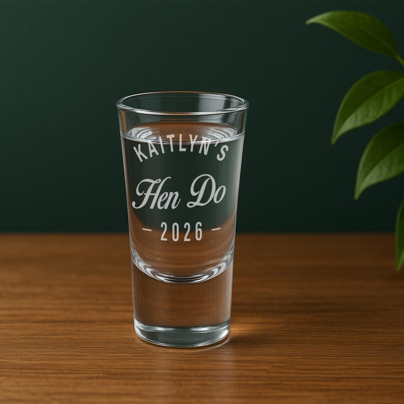 May include: A clear shot glass filled with liquid, featuring the text "Kaitlyn's Hen Do - 2026" in a decorative font. The glass is set on a wooden surface against a dark green background, with a plant visible on the right.