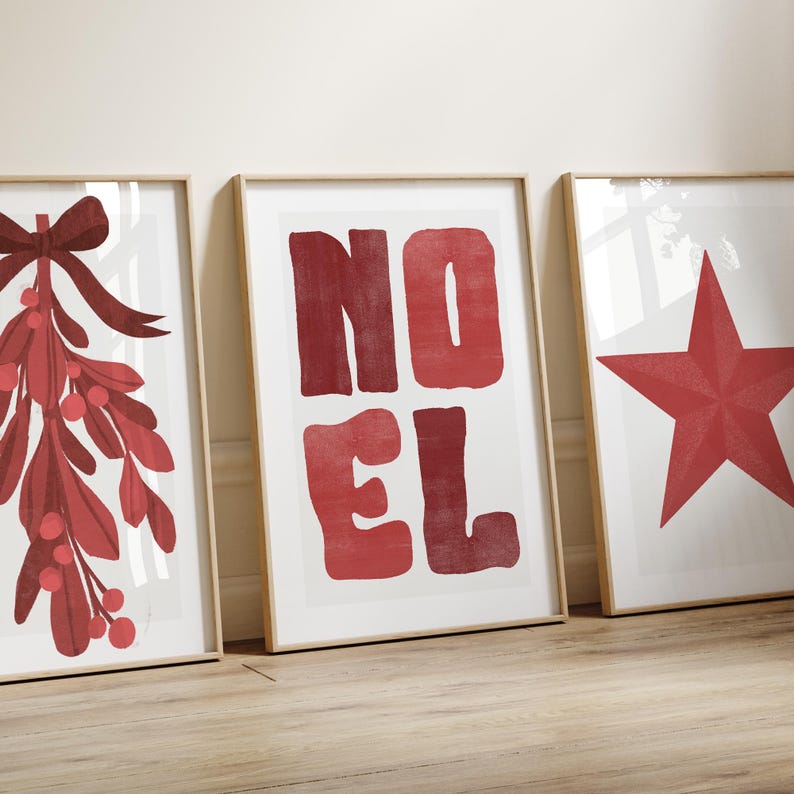 May include: Three framed art prints with a Christmas theme. The prints feature a red mistletoe with a bow, the word "NOEL" in red block letters, and a red star. The frames are a light wood colour.