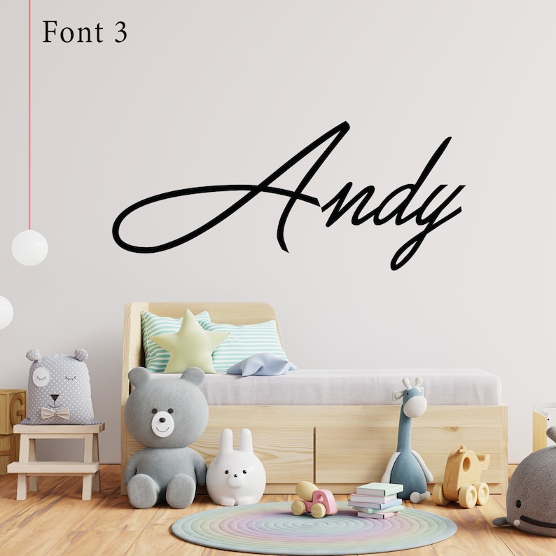 Op de afbeelding: Een witte muur met de naam "Andy" geschreven in een zwart cursief lettertype. De muur bevindt zich achter een houten bed met een lichtblauw en wit gestreept kussen, een lichtgroen stervormig kussen en een grijs knuffeldier.
