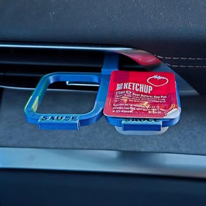 May include: A blue plastic car vent holder with two compartments. The holder is labeled "SAUCE". A red ketchup packet is in one compartment. The packet is labeled "KETCHUP" and has a tomato image. The text "21ml Best Before: See Pot" is printed on the packet.