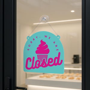 Może przedstawiać: Turkusowy znak z r&oacute;żową grafiką babeczki i napisem "Closed" w kursywie. Znak wisi na szklanych drzwiach, wskazując, że firma jest nieczynna.