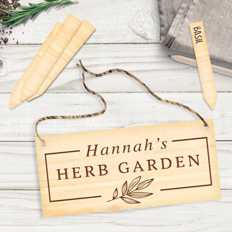 May include: A wooden sign with the text "Hannah's Herb Garden" in brown lettering. The sign is hanging from a twine rope and has a small leaf design. There are three wooden plant markers and a cloth napkin with the word "Basil" written on it.