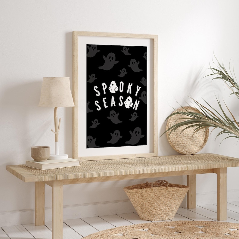 Puede incluir: Una impresi&oacute;n art&iacute;stica enmarcada con un fondo negro y texto blanco que dice "Spooky Season". La impresi&oacute;n presenta un patr&oacute;n de ilustraciones de fantasmas grises. El marco es de color madera clara y la impresi&oacute;n se exhibe en un banco de madera clara.