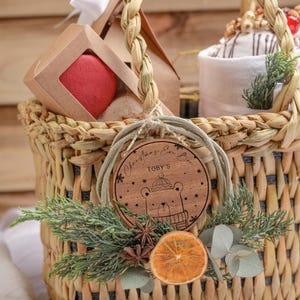 May include: A wicker basket filled with gifts for the festive season. The basket is decorated with a wooden ornament that says "Christmas Eve Box Toby's" and features a cartoon bear wearing a hat. The basket also includes a red macaron, a white towel, and a dried orange slice.