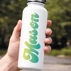 May include: White water bottle with a silver lid, featuring the name "Mason" in a gradient of yellow and teal. The bottle is held by a hand, with a blurred green background.