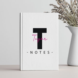 Pode incluir: Caderno branco com um T mai&uacute;sculo preto e a palavra "Taylor" escrita em it&aacute;lico rosa acima. As palavras "NOTES" est&atilde;o escritas em preto abaixo da letra T.