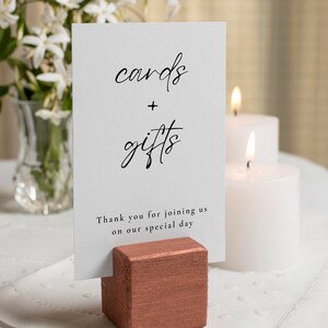 May include: A white sign with the words "cards + gifts" in elegant black script, held by a copper-coloured wooden block. The sign also includes the text "Thank you for joining us on our special day". Two white candles are lit in the background.