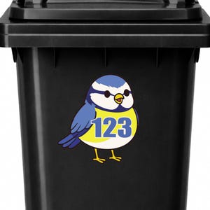 Op de afbeelding: Zwarte vuilnisbak met een blauwe en gele vogelsticker. De vogel heeft het nummer 123 op zijn buik. De vogel heeft blauwe vleugels en een witte kop met een blauwe pet. De vogel heeft gele poten en een gele snavel.