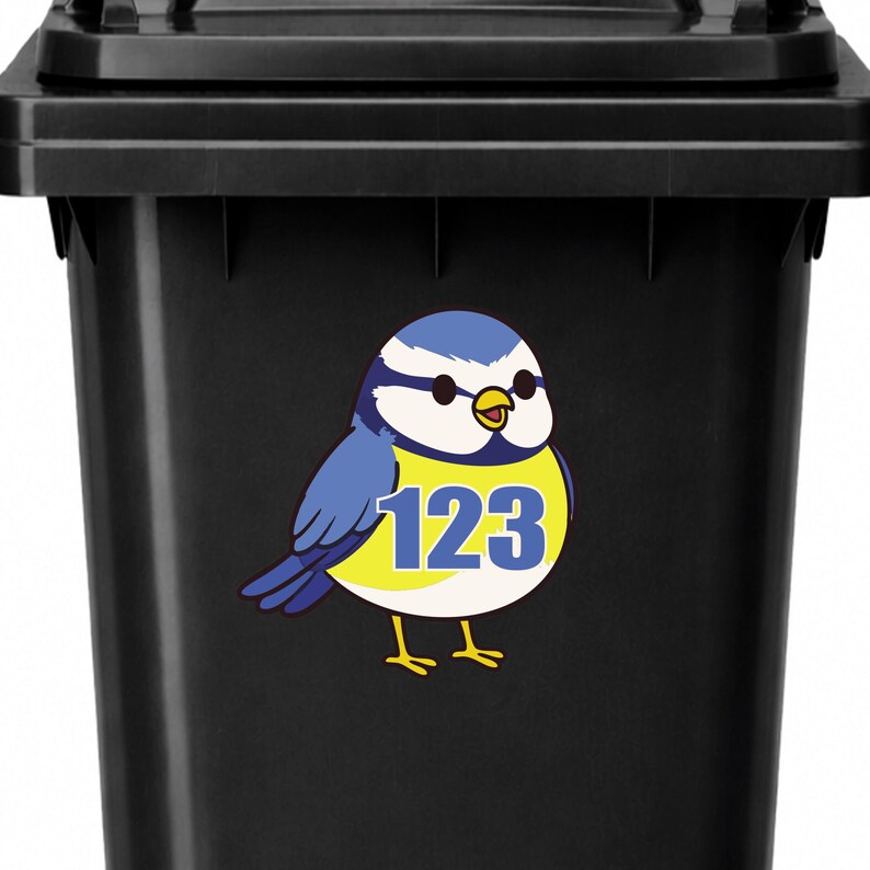 Op de afbeelding: Zwarte vuilnisbak met een blauwe en gele vogelsticker. De vogel heeft het nummer 123 op zijn buik. De vogel heeft blauwe vleugels en een witte kop met een blauwe pet. De vogel heeft gele poten en een gele snavel.