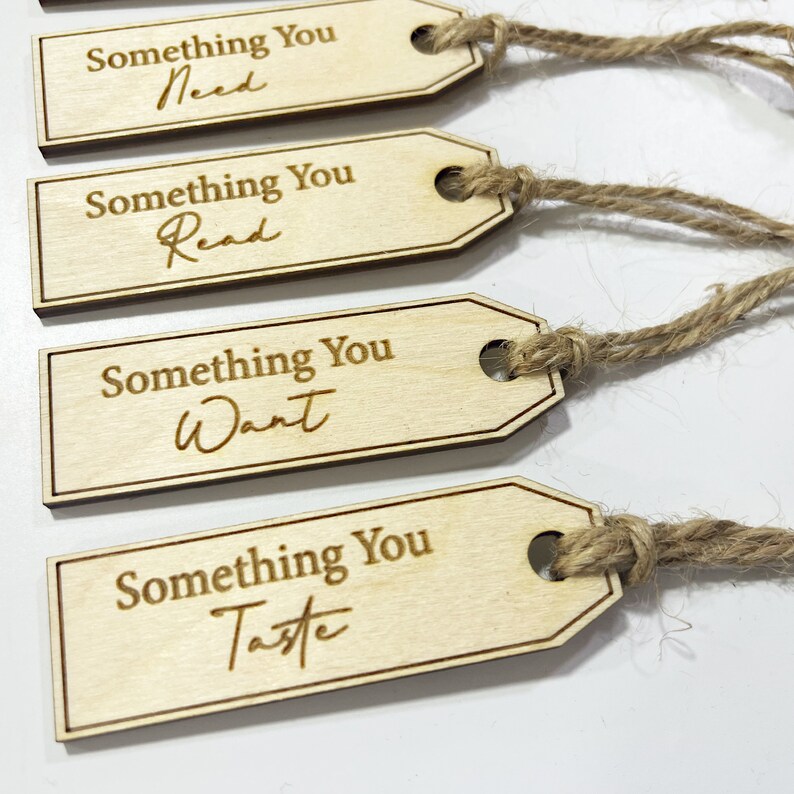 Pu&ograve; includere: Quattro etichette regalo in legno con spago attaccato. Ogni etichetta ha il testo "Something You" seguito da una parola diversa: "Need", "Read", "Want" e "Taste".