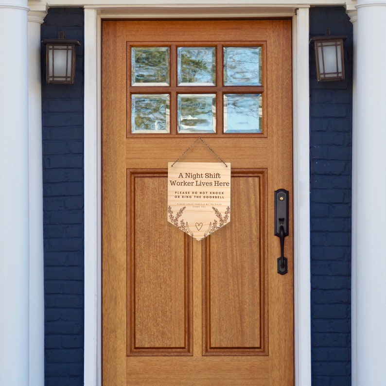 May include: Wooden sign hanging on a wooden door. The sign reads "A Night Shift Worker Lives Here. Please do not knock or ring the doorbell." The sign has a heart and floral design.