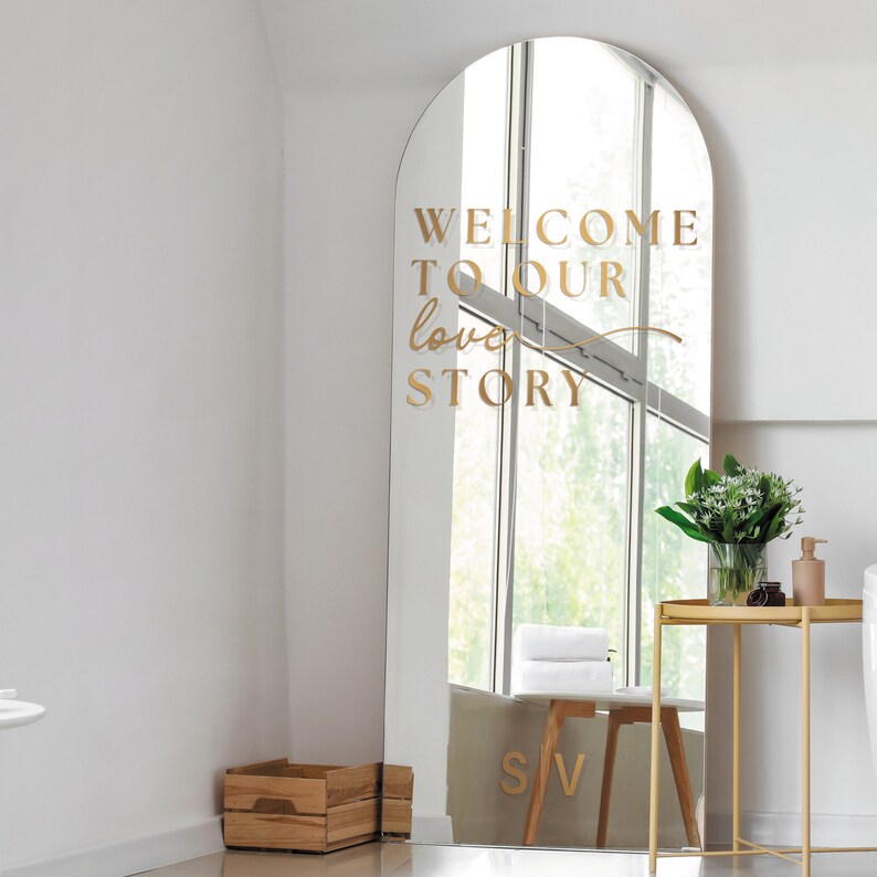 May include: A large, arched mirror with gold lettering that reads "WELCOME TO OUR love STORY." The mirror is in a bright room with a window, a small table, and a wooden crate. The mirror is a decorative piece.