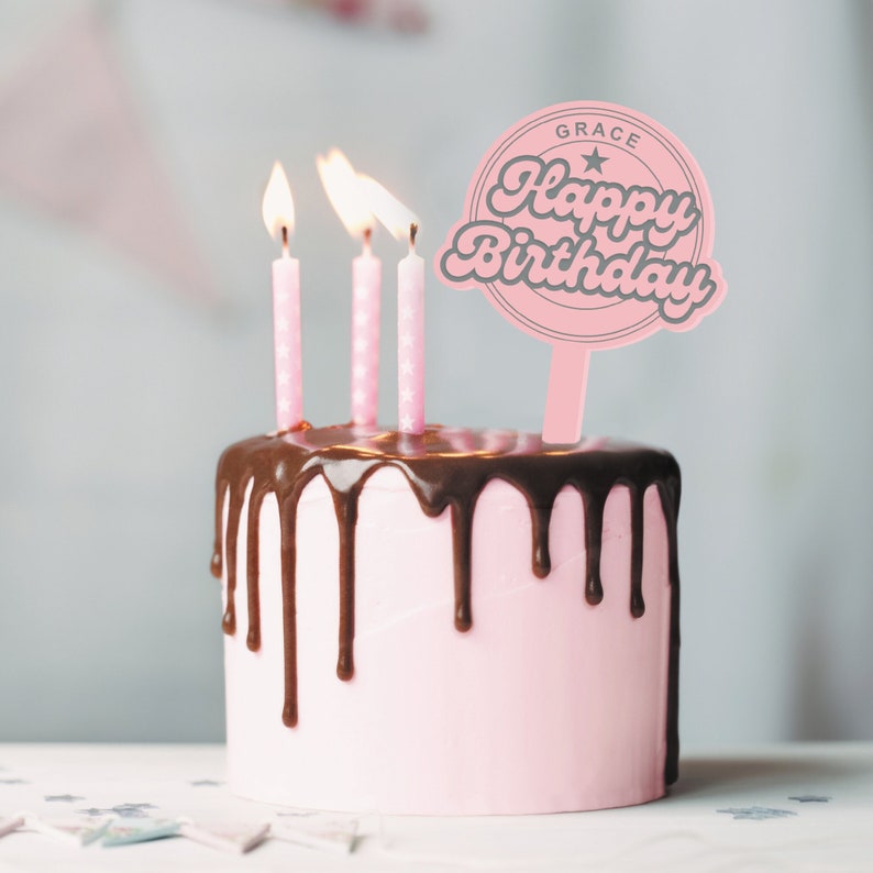 May include: A pink cake with chocolate frosting and three lit candles. A pink cake topper with the text "GRACE Happy Birthday" is on top of the cake.