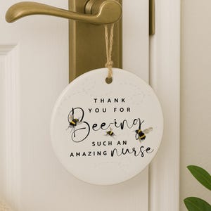 May include: A round, white ceramic door hanger with a bee theme. The text reads "Thank You For Bee-ing Such An Amazing Nurse," with bee illustrations. It hangs from a door handle with a natural twine.