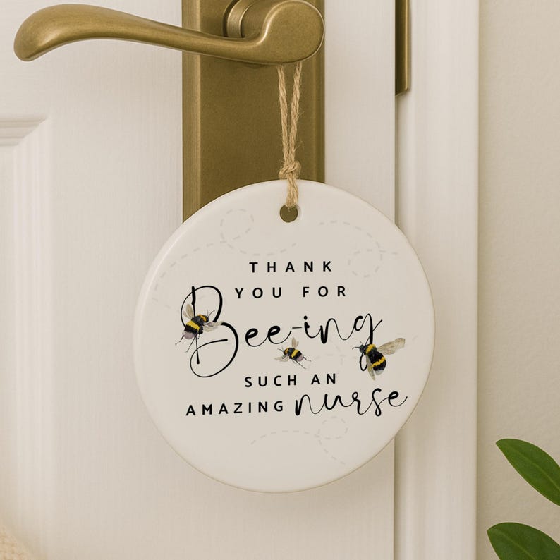 May include: A round, white ceramic door hanger with a bee theme. The text reads "Thank You For Bee-ing Such An Amazing Nurse," with bee illustrations. It hangs from a door handle with a natural twine.