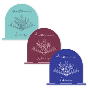 May include: Three bookends in different colors: teal, burgundy, and blue. Each bookend features a white outline of an open book with flowers growing from it. The text "Anastasia's Library" is written on each bookend.