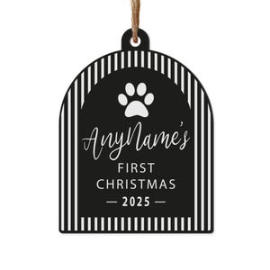 May include: A black and white pet-themed Christmas ornament. The ornament has a paw print and the text "AnyName's FIRST CHRISTMAS - 2025". The ornament is rectangular with a rounded top and a striped border, and is suspended by a brown string.