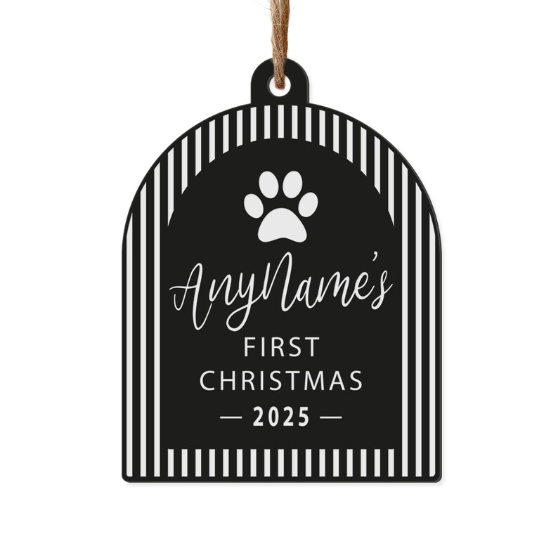 May include: A black and white pet-themed Christmas ornament. The ornament has a paw print and the text "AnyName's FIRST CHRISTMAS - 2025". The ornament is rectangular with a rounded top and a striped border, and is suspended by a brown string.
