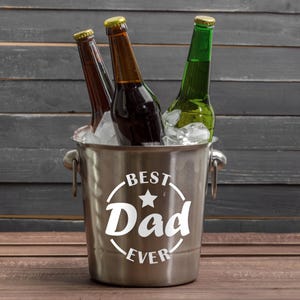 Puede incluir: Un cubo de hielo plateado con el texto "Best Dad Ever" en letras blancas y una estrella. Tres botellas de cerveza est&aacute;n dentro del cubo con hielo.
