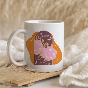 May include: White ceramic mug with a graphic of two dice with hearts on them, flames, and stars. The text on the mug reads "I'm lucky to have you."
