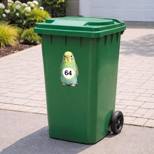 May include: A green outdoor trash can with a matching lid and black wheels. A sticker of a colorful parakeet with the number 64 is affixed to the front of the can. The can is on a paved surface with greenery in the background.