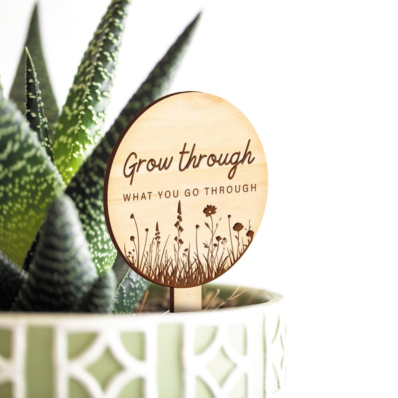 K&ouml;nnte beinhalten: Ein rundes Holzschild mit dem Text "Grow through what you go through" und einer Silhouette von Wildblumen. Das Schild ist an einem Holzstab befestigt.