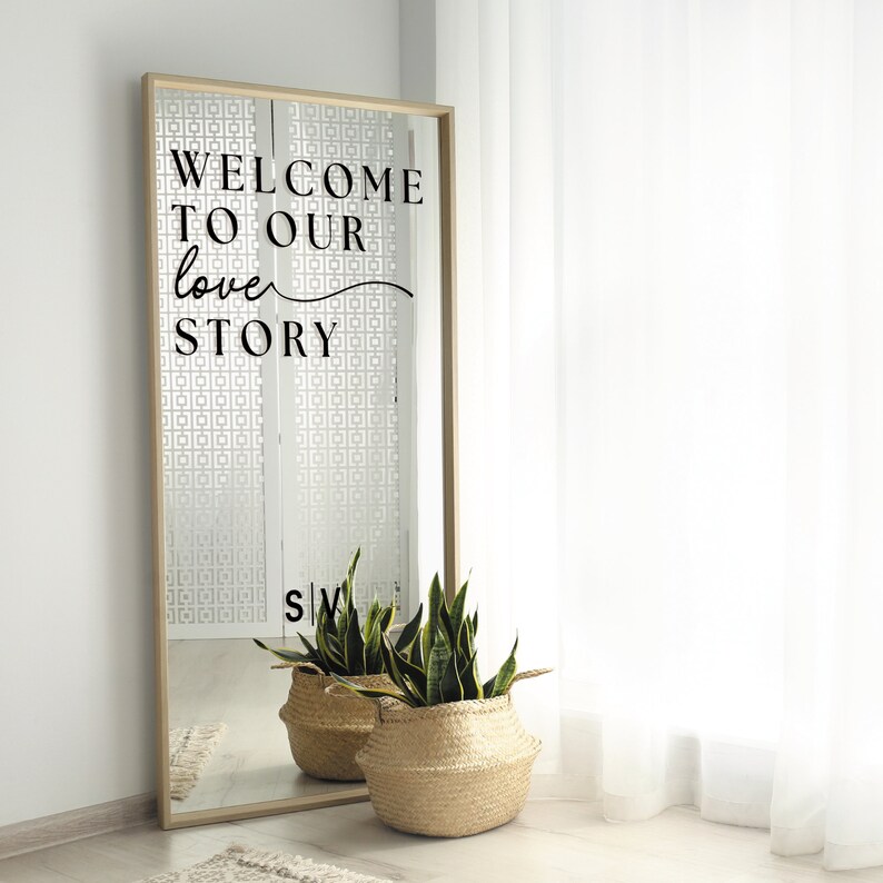 May include: A full-length mirror with a wooden frame. The mirror has the text "WELCOME TO OUR love STORY" in black. Two woven baskets with green plants sit in front of the mirror.
