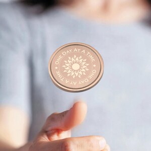 May include: A round, copper-colored token with the words "One Day At A Time" and a stylized sun design.