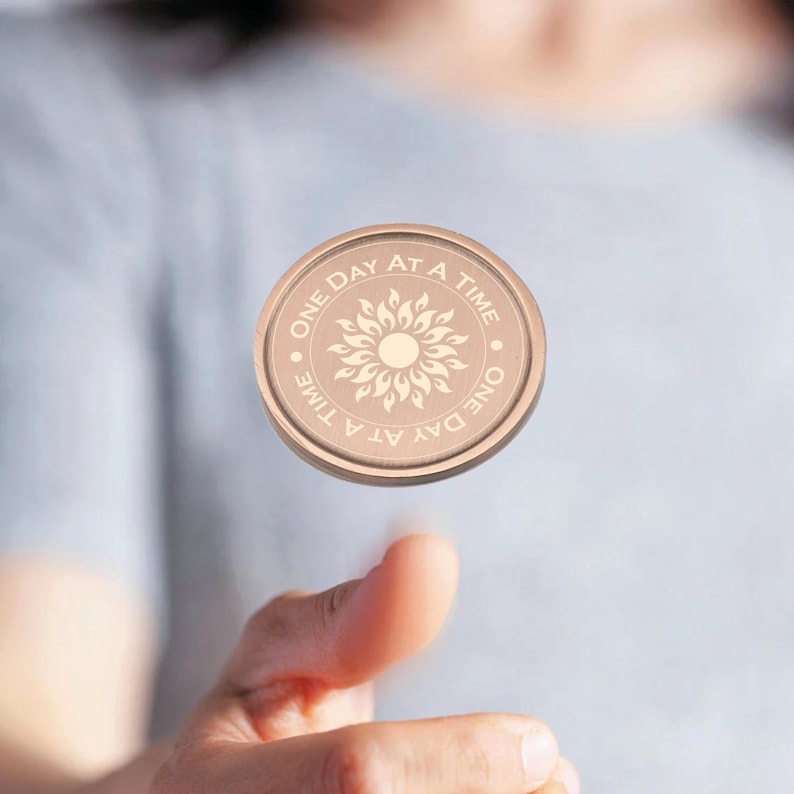 May include: A round, copper-colored token with the words "One Day At A Time" and a stylized sun design.