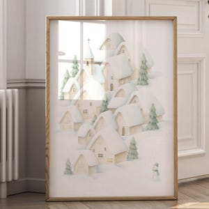 May include: A framed print showing a watercolour illustration of a snow-covered village. The artwork depicts houses with snow-laden roofs, evergreen trees, and a small snowman. The frame is made of light-coloured wood.