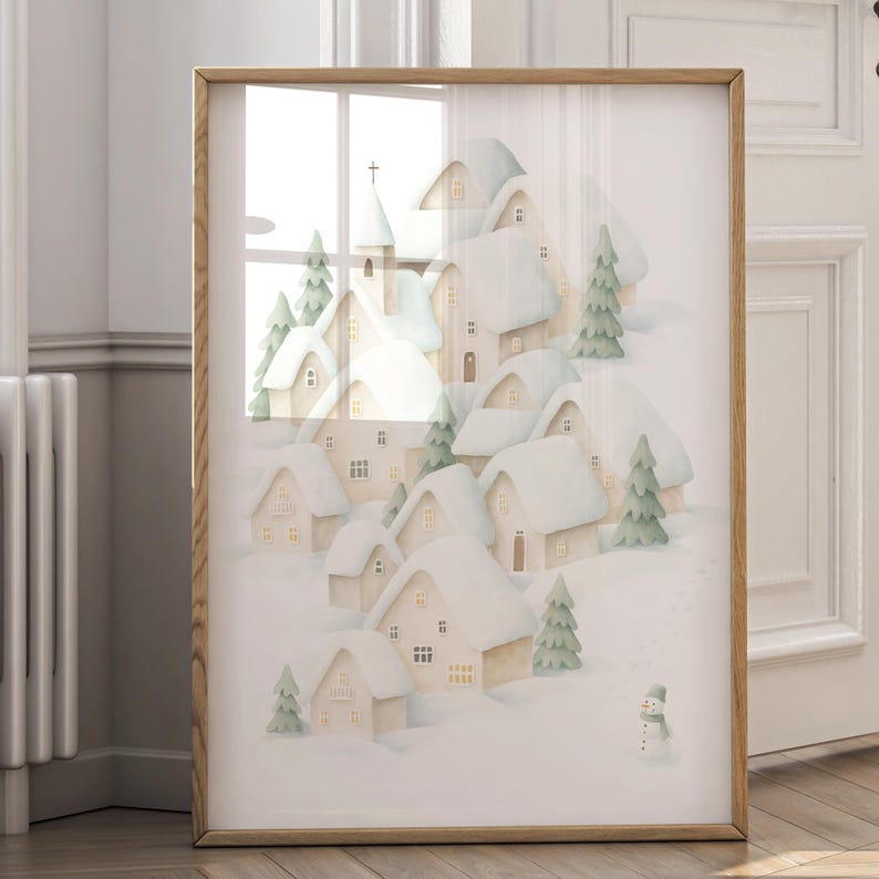 May include: A framed print showing a watercolour illustration of a snow-covered village. The artwork depicts houses with snow-laden roofs, evergreen trees, and a small snowman. The frame is made of light-coloured wood.