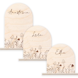 May include: Three wooden arch-shaped signs with the names Anastasia, Lola, and Ellie written in cursive. Each sign has a decorative border of mushrooms and flowers.