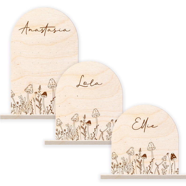 May include: Three wooden arch-shaped signs with the names Anastasia, Lola, and Ellie written in cursive. Each sign has a decorative border of mushrooms and flowers.