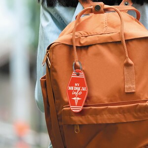 May include: A red motel key tag with white text that says "MY MEDICAL info" and a white medical symbol. The key tag is attached to a brown backpack.