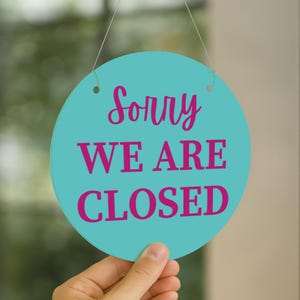 K&ouml;nnte beinhalten: Ein rundes, t&uuml;rkisfarbenes Schild mit dem Text "Sorry WE ARE CLOSED" in rosa Schrift. Das Schild h&auml;ngt an einer Schnur und wird von einer Hand gehalten. Der Hintergrund ist unscharf.