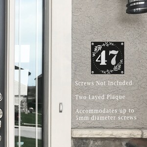 May include: Black and white house number plaque with the number 47 in a white circle surrounded by a floral design. The plaque is mounted on a gray wall. The text "Screws Not Included", "Two Layered Plaque", and "Accommodates up to 5mm diameter screws" is printed below the plaque.