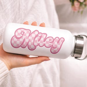 May include: White water bottle with a silver lid and handle. The bottle features the name "Miley" in a pink checkered font. The bottle is held by a person.