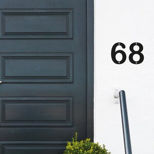 May include: Black metal house numbers, "68", are fixed to a white wall with small silver screws. A dark teal door is on the left, and a small green bush is at the bottom. A black handrail is on the right.