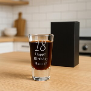 May include: A clear shot glass filled with a dark liquid, featuring the etched text "18 Happy Birthday Hannah." A black gift box is behind the glass. The shot glass is on a light wood surface.