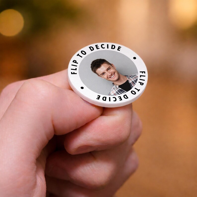 May include: A white, circular decision coin with the text "FLIP TO DECIDE" in black around the edge. The coin features a photo of a smiling person. The coin is held in a hand.
