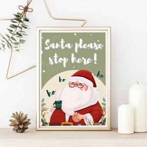 May include: A printable sign featuring a cartoon Santa Claus holding a cup of hot cocoa and a cookie. The sign says "Santa please stop here!" with "ho ho ho" written around Santa.
