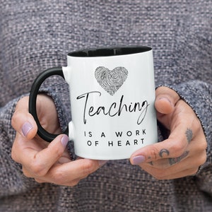 Puede incluir: Taza de cer&aacute;mica blanca con asa e interior negros. La taza presenta el texto "Teaching is a work of heart" y un dise&ntilde;o de coraz&oacute;n de huellas dactilares. Un regalo para profesores.