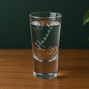 以下が含まれることがあります： 筆記体のフォントで「Hannah」という名前と数字の「30」が刻印された、透明なガラス製のショットグラス。厚みのある底と円筒形の形状をしており、少量の液体を入れるために設計されています。