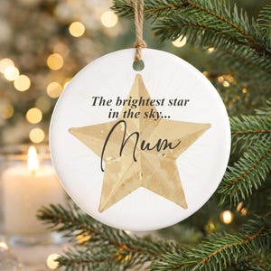 May include: A white ceramic Christmas ornament with a gold star design and the word "Mum" in cursive. The ornament has the text "The brightest star in the sky..." and hangs from a natural twine string. The ornament is displayed on a Christmas tree.