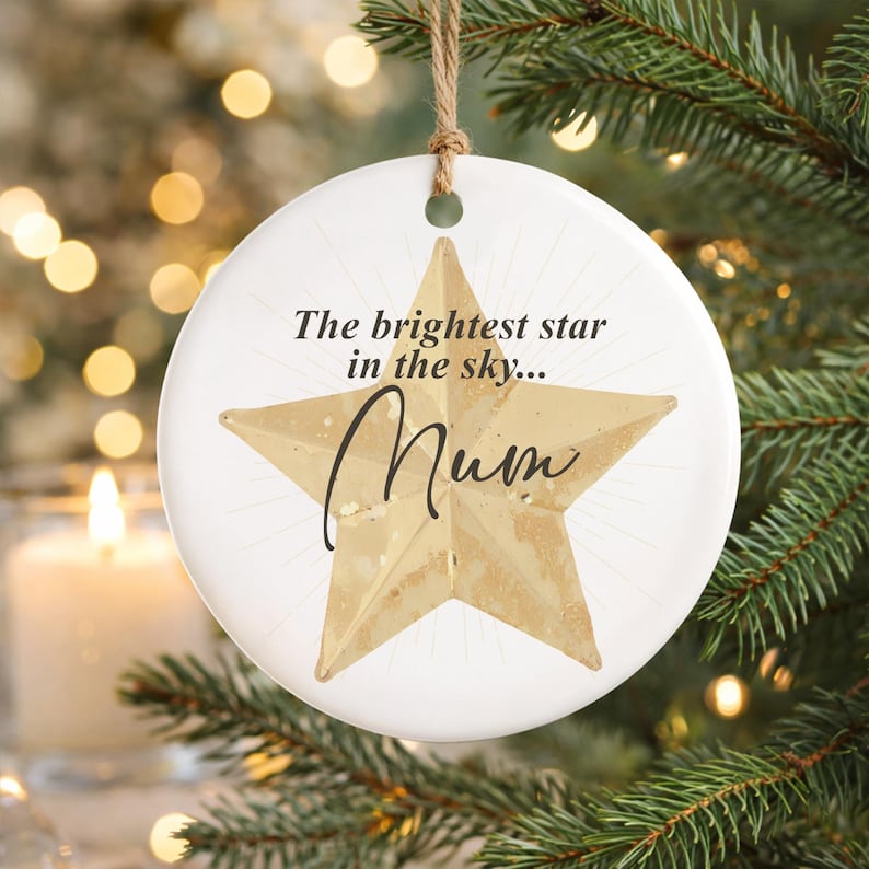 May include: A white ceramic Christmas ornament with a gold star design and the word "Mum" in cursive. The ornament has the text "The brightest star in the sky..." and hangs from a natural twine string. The ornament is displayed on a Christmas tree.