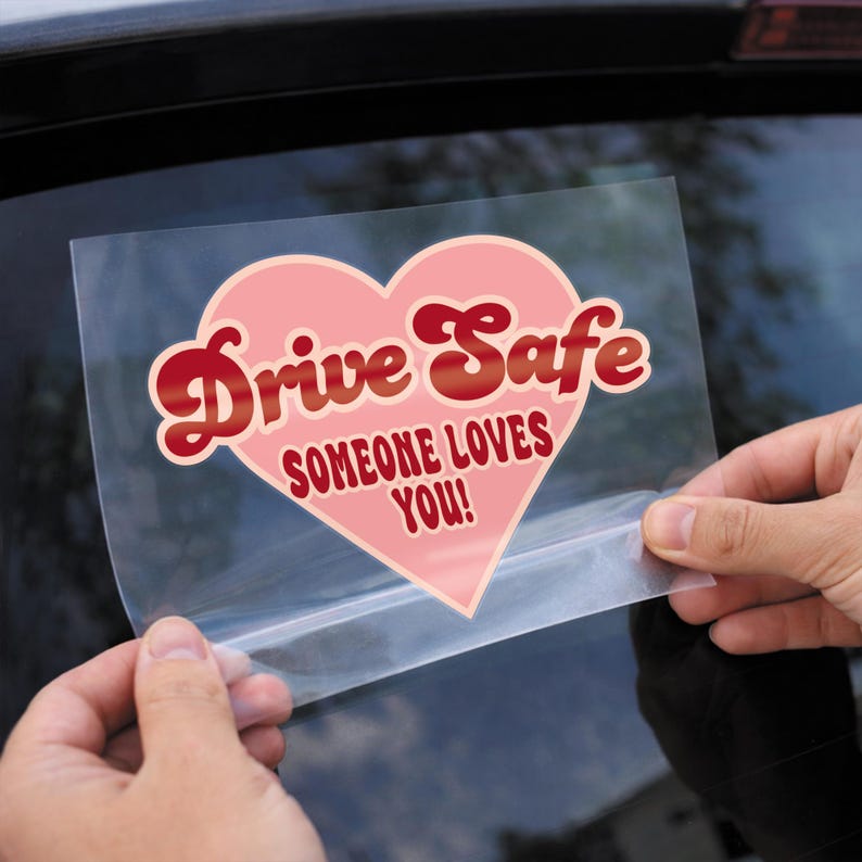 May include: A heart-shaped car decal with the words "Drive Safe" in red script over the words "Someone Loves You!" in a pink heart. The decal is held up against a car window.