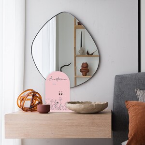May include: A pink earring display stand with the name Anastasia printed on it. The stand features a floral design with mushrooms and butterflies. The stand is on a wooden table with a brown bowl and a brown decorative object.