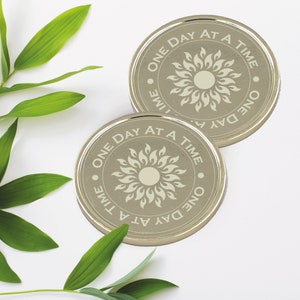 May include: Two silver-colored round coasters with the phrase "One Day At A Time" and a stylized sun design.