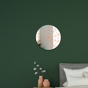 May include: A round mirror with an orange spiderweb design. The mirror is hanging on a dark green wall above a bed with white bedding.